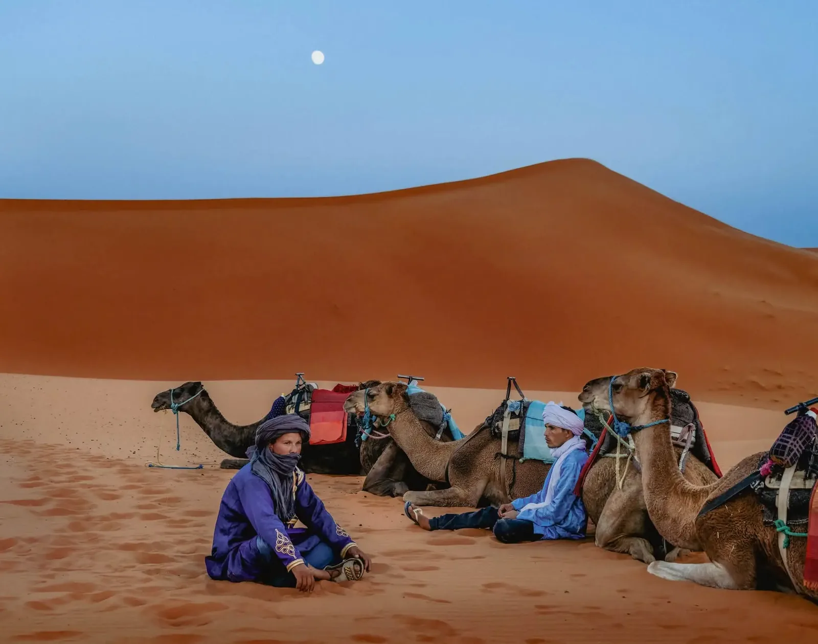 Deux hommes en tenue traditionnelle assis près de leurs dromadaires au pied d’une grande dune